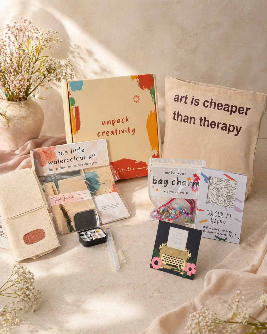 Collection of art supplies and decorative items on a table with a 'unpack creativity' book and 'art is cheaper than therapy' pillow.